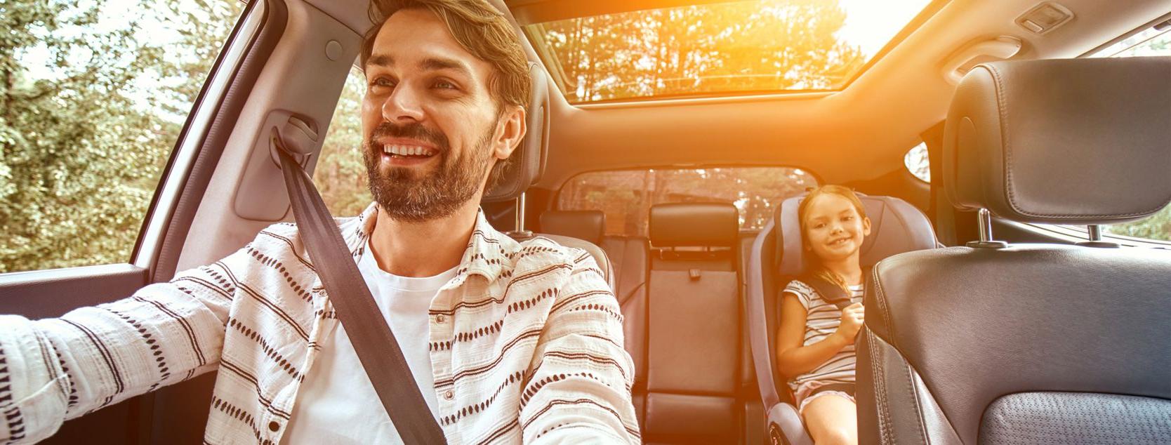 A smiling dad talks to his daughter as they drive the family car through a wooded area.